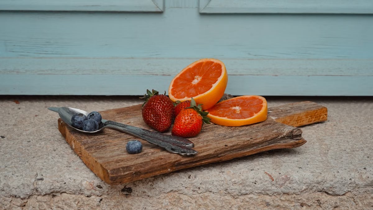 A rustic display of strawberries, oranges, and blueberries on a wooden board against a blue backdrop.